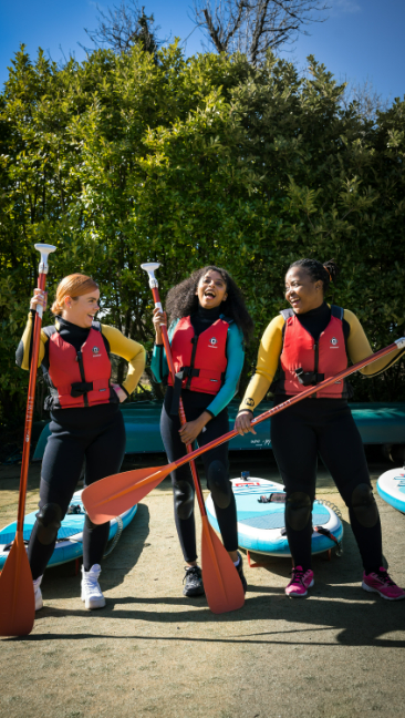 Three women in wetsuits and life jackets, with oars and paddleboards behind them