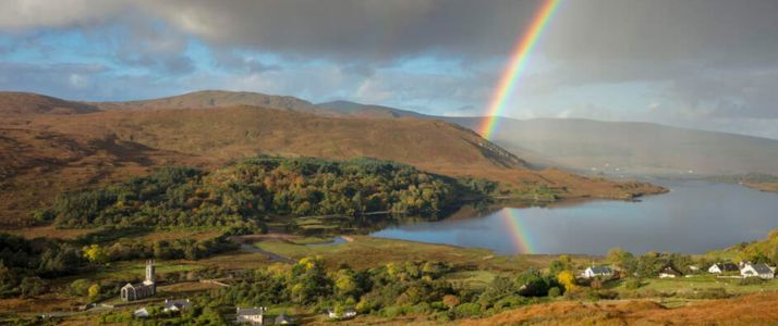 View of a small town by a lake, there is a mountain in the distance. It is a bit cloudy, but there is some sun as a rainbow crosses over the right hand side of the image into the bottom of the mountain.