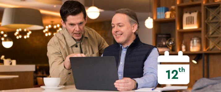 Two men looking at a laptop with a calendar icon that reads the 12th